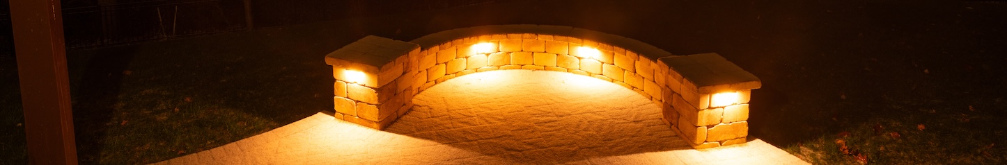A backyard patio with a curved stone wall glowing under warm lights at night. The dark forest and distant house lights create a peaceful and cozy evening atmosphere.