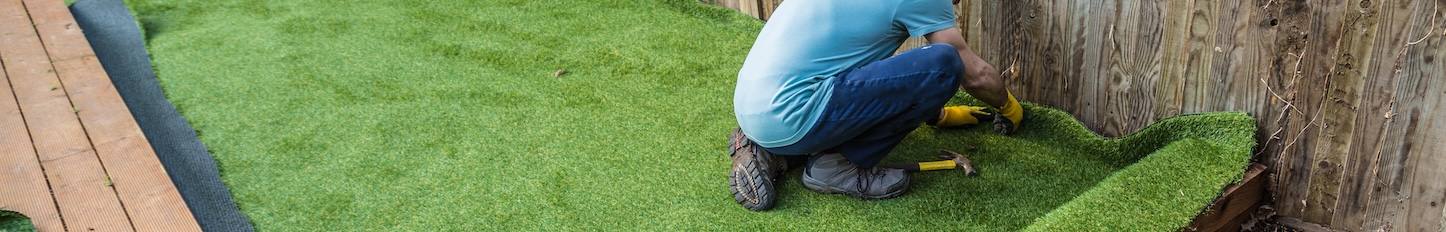 Artificial grass being installed, added next to wooden decking. It has been cut to size and rolled out and laid and is being nailed down. the installer has a hammer and nails.